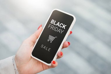 female hand holding mobile phone with black friday text and shopping cart of the background of a street clipart