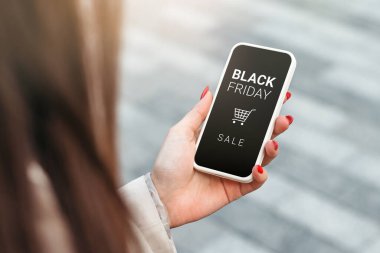 Elinde siyah cuma mesajları ve arka plandaki alışveriş arabasıyla akıllı telefon tutan bir kadın.