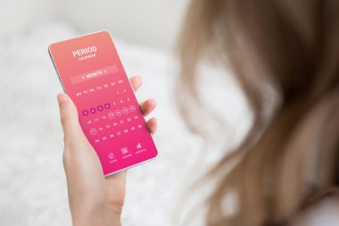 Elinde adet döngüsü takvimi olan cep telefonu tutan ve odasında oturan bir kadın.