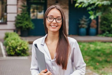 Gözlüklü öğrenci bir klasör tutuyor ve binanın ön cephesine gülümsüyor. Uzaktan eğitim. Yurtdışında eğitim