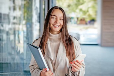 Akıllı telefon ve klasör tutan genç bir kadın bir ofis binasının arka planında kameraya bakıyor. Boşluğu kopyala