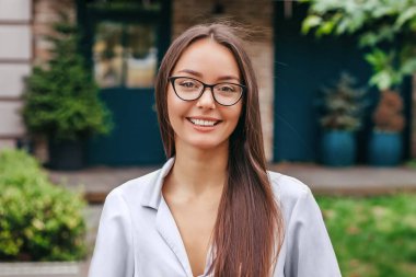 Gözlüklü öğrenci bir klasör tutuyor ve binanın ön cephesine gülümsüyor. Uzaktan eğitim. Yurtdışında eğitim