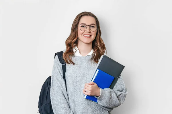 Sırt çantalı ve gözlüklü bir öğrenci kız gülümsüyor ve elinde beyaz bir arka planda defter, defter ve klasör tutuyor.