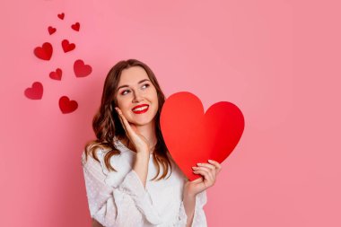 Girl in love makes a wish and gift for valentines day isolated on pink background. Young woman smiling and thoughtfully holding a valentine card