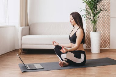 Güzel bir kadın evde yoga yapıyor ve güneşli bir günde dizüstü bilgisayarda ders izliyor. Kız evde meditasyon yapmayı, nefes almayı öğreniyor.