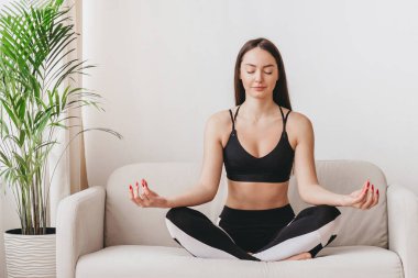 Kafkasyalı kız gözleri nilüfer pozisyonunda kanepede oturur ve meditasyon yapar. Akıl sağlığı, nefes alma pratiği, duygu kontrolü, evde yoga.