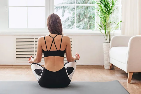 Yoga and meditation at home. Back view of a young woman in sportswear sitting in lotus position doing yoga breathing practices