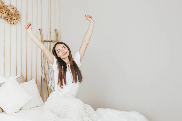 stock image Happy morning concept. yound woman sitting on the bed, stretching after sleep. Attractive young female in a white t-shirt warm up before workout, copy space