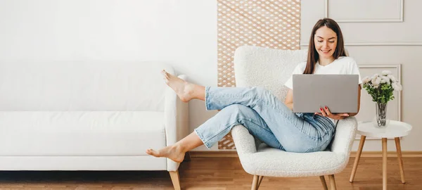 stock image interior designer web banner. young woman using laptop at home. brunette woman sitting on chair smiling and look on computer, wear white shirt and jeans. Freelance concept. copy space