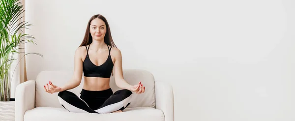 年轻的女人坐在沙发上 眼睛紧闭在荷花的位置上沉思 精神健康 呼吸练习 情绪控制 瑜伽在家 网页横幅 复制空间 — 图库照片
