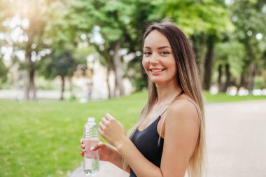 Mutlu gülümseyen kız elinde bir şişe suyla parkta koşuya çıktı. Spor ve sağlıklı yaşam tarzı