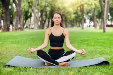 Genç esmer, beyaz kadın parkta egzersiz yaparken yoga yapıyor. Sağlıklı bir yaşam tarzı. Doğada fiziksel egzersiz. Kız parkta meditasyon yapıyor.