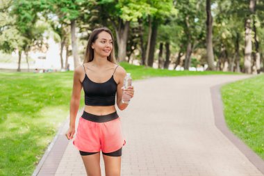Parkta bir şişe suyla sabah koşusunda olan mutlu genç bir kadın. Spor ve sağlıklı yaşam tarzı. Boşluğu kopyala