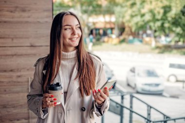 Mutlu kız öğrenci bir cep telefonu ve bir fincan kahve tutuyor ve üniversite binasının yanında duruyor. Boşluğu kopyala