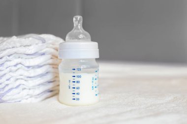 Beyaz kumaş arka planında süt ve bebek bezi olan bebek şişesi. Bebek bakımı. Hayatın ilk günleri. Beslenme ve annelik. Boşluğu kopyala