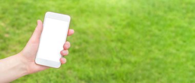 Bir kadın eli, yeşil çimenlerin arka planında boş ekranlı beyaz bir akıllı telefonu tutuyor. Modelleme. Uzayı kopyala Web pankartı