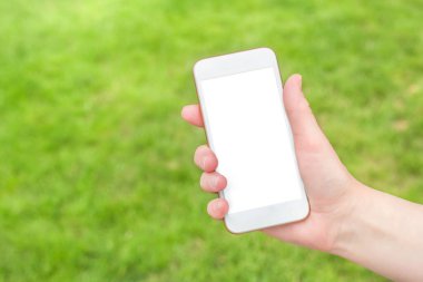 Bir kadın eli, yeşil çimenlerin arka planında boş ekranlı beyaz bir akıllı telefonu tutuyor. Modelleme. Uzayı kopyala Tasarım için yer