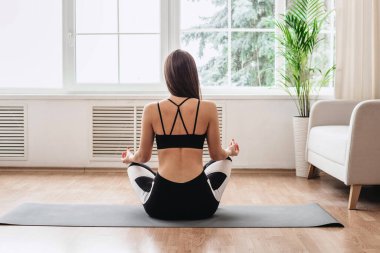 Yoga ve evde meditasyon. Nilüfer pozisyonunda oturmuş yoga yapan spor giysili narin genç bir kadının arka görüntüsü. Huzurlu kız evde nilüfer pozisyonunda dinleniyor yoga minderinde pencerelere bakıyor..