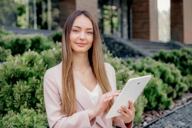 İş elbisesi giymiş beyaz bir kadın elinde tabletle bir ofis binasının arka planındaki çiçek tarhlı kameraya bakıyor.