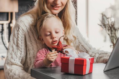 Küçük bir oğlu olan anne oyuncaklara ve hediyelere bakıyor ve mutfak masasında pencerenin arkasında oturuyor.