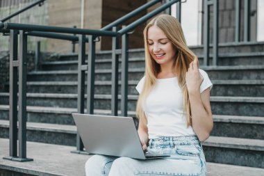 Üniversitenin yanındaki merdivenlerde dizüstü bilgisayarı olan genç bir kız gülümsüyor ve monitöre bakıyor.