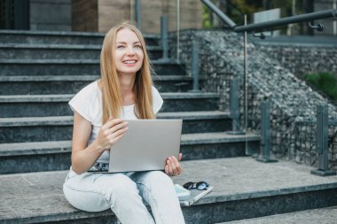 Üniversitenin yanındaki merdivenlerde dizüstü bilgisayarla oturan genç kız gülümsüyor ve çalışıyor.