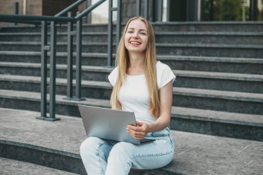 Üniversitenin yanındaki merdivenlerde dizüstü bilgisayarlı genç beyaz kız gülümsüyor ve çalışıyor.