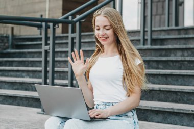 Üniversitenin yanındaki merdivenlerde dizüstü bilgisayarla oturan genç kız gülümsüyor ve çalışıyor. video çağrıları konsepti