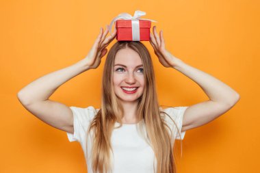 Mutlu, beyaz, genç, sarışın bir kadın başının üstünde kırmızı bir hediye kutusu tutuyor. Stüdyoda sarı arka planda izole edilmiş.