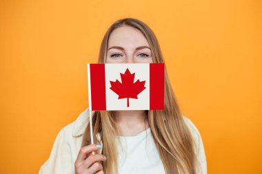 Kafkasyalı kız öğrencinin portresi, yüzünü küçük Kanada bayrağıyla örtüyor ve turuncu arka planda izole edilmiş kameraya bakıyor, Kanada günü, bayram, konfederasyon yıldönümü, kopyalama alanı