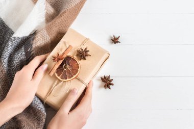 Kadın eli, anason, kuru koni, turuncu ve tarçın kaplı bir hediye kutusunu taşra tarzı beyaz ahşap arka planda tutuyor. Kopya alanı, üst görünüm.