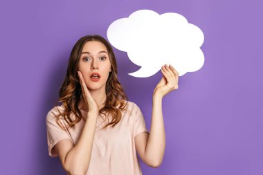 Genç, şok olmuş bir kız öğrenci elinde mor arka planda izole edilmiş boş konuşma balonu kağıdı tutuyor. Leylak üzerine yazı için beyaz kağıt bulutu. Boşluğu kopyala
