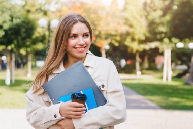 Sarışın kız öğrenci elinde klasörler, not defterleri ve bir fincan kahve, eğitim konseptiyle parkta yürüyor.