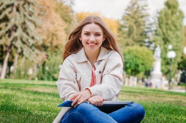 Çimlerin üzerinde oturan sarışın kız öğrenci ve not defterine notlar alıyor. Öğrenci kız gülümser ve ders verir.