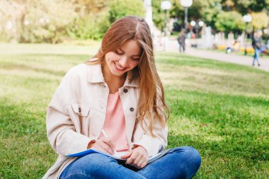 Çimlerin üzerinde oturan sarışın kız öğrenci ve not defterine notlar alıyor. Öğrenci kız defterine bakar ve yazar.