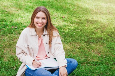 Sarışın kız öğrenci çimlerin üzerinde oturuyor ve not defterine not alıyor, fotokopi odası. Öğrenci kız gülümser ve ders verir.