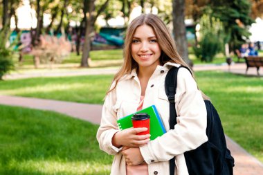 Sarışın kız öğrenci elinde bir defter ve bir fincan kahveyle parkta yürüyor. Eğitim kavramı