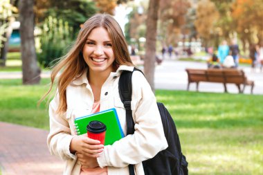 Sarışın kız öğrenci gülüyor ve elinde defterler ve bir fincan kahveyle parkta yürüyor. Eğitim kavramı