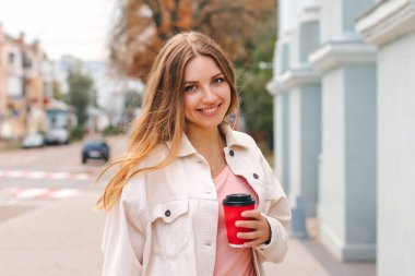 Şirin sarışın kız bir fincan kahveyle şehirde yürüyor ve gülümsüyor.