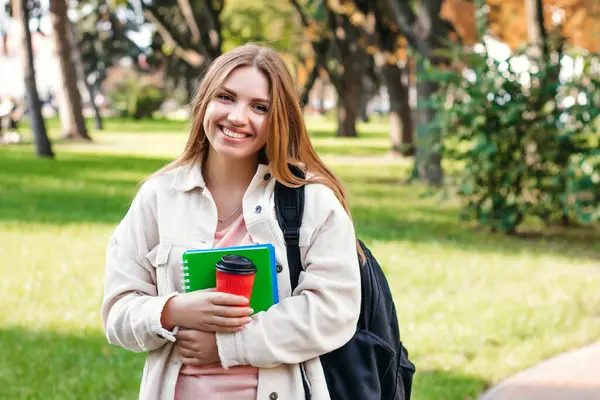 Sarışın kız öğrenci elinde bir defter ve bir fincan kahveyle parkta yürüyor. Eğitim kavramı