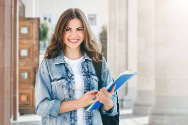 Üniversiteye karşı gülümseyen genç bir kız. Sevimli kız öğrenci klasörleri ve defterleri elinde tutuyor. Öğrenme, eğitim kavramı