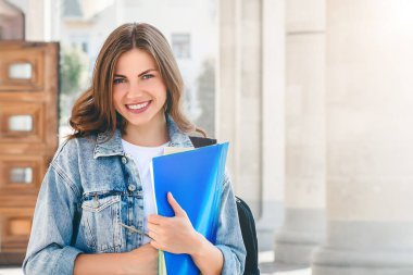 Üniversiteye karşı gülümseyen genç bir kız. Sevimli kız öğrenci klasörleri ve defterleri elinde tutuyor. Öğrenme, eğitim kavramı