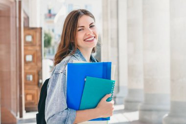 Üniversiteye karşı gülümseyen genç bir kız. Sevimli kız öğrenci klasörleri ve defterleri elinde tutuyor. Öğrenme, eğitim kavramı