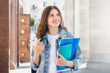 Üniversiteye karşı gülümseyen genç bir kız. Sevimli kız öğrenci klasörleri, defterleri ve cep telefonunu elinde tutuyor. Öğrenme, eğitim kavramı