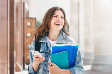 Üniversiteye karşı gülümseyen genç bir kız. Sevimli kız öğrenci klasörleri, defterleri ve cep telefonunu elinde tutuyor. Öğrenme, eğitim kavramı