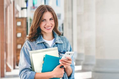 Üniversiteye karşı gülümseyen genç bir kız. Sevimli kız öğrenci klasörleri, defterleri ve cep telefonunu elinde tutuyor. Öğrenme, eğitim kavramı