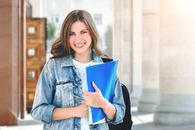Üniversiteye karşı gülümseyen genç bir kız. Sevimli kız öğrenci klasörleri ve defterleri elinde tutuyor. Öğrenme, eğitim kavramı