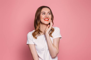 Kırmızı rujlu, beyaz tişörtlü, beyaz tişörtlü genç beyaz bir kadın pembe arka planda yanak kenarında fotokopi odasına bakıp el ele tutuşuyor.