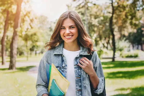 Parkta gülümseyen genç kız öğrenci. Sevimli kız öğrenci klasörleri ve defterleri elinde tutuyor. Öğrenme, eğitim kavramı