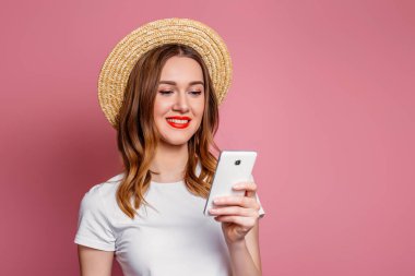 Hasır şapkalı, beyaz tişörtlü, kırmızı rujlu genç bir kız gülümsüyor ve elinde bir cep telefonu tutuyor. Kadın akıllı telefon kullanıyor ve SMS mesajı okuyor